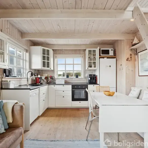 Et lyst køkken i træ med hvide skabe og sorte bordplader. En stige fører op til en hems. En sofa og bord står ved siden af, under et vindue med udsigt.