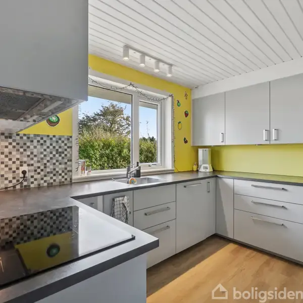 Køkken med grå skabe og skuffer, flisebelagt væg, og sort kogeplade; vindue åbent til have med buske og træer udenfor; gule vægge og trægulv.