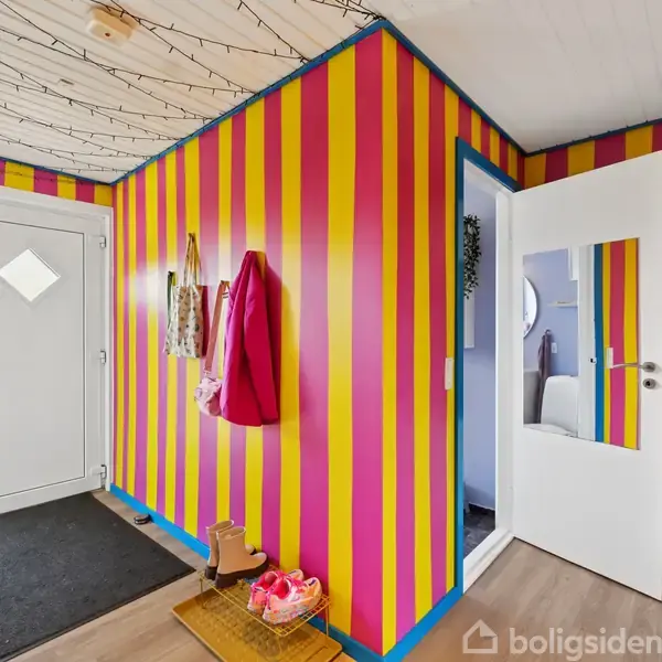 Entré med farverige stribede vægge i gul, pink og blå. Jakker hænger på knager, og et par sko står på en skohylde. Døren til et rum står på klem.