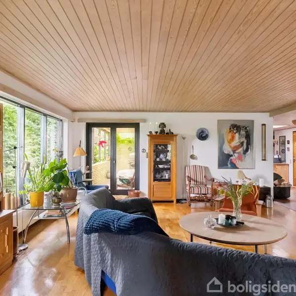 Stue med træloft og trægulv. Møbler omfatter hylder med bøger, sofaer og et rundt sofabord. Store vinduer viser grønne planter udenfor. Væggen har kunstmaleri og enkel dekoration.