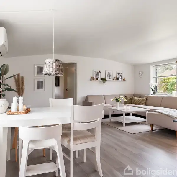 Stue med et spisebord og fire stole i forgrunden. Sofa med sofabord i baggrunden. Væggene har reoler med dekorationer. Lyseffekter fra store vinduer skaber et lyst og indbydende rum.