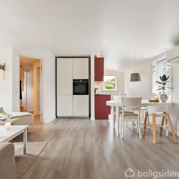 Stue og køkken i et åbent rum med lyst trægulv. Sofa og sofabord i venstre side, spisebord og stole til højre. Aircondition på væggen over spisebordet.