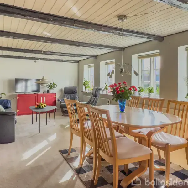 Et spisebord med træstole er centralt placeret i en lys stue. Der står en vase med blomster på bordet, omgivet af vinduer og en sofa.