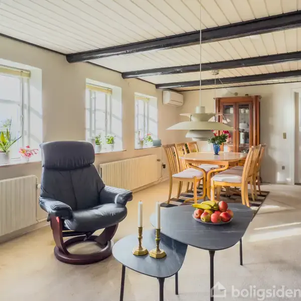 Stue med lænestol og sofabord med frugt og lys på. I baggrunden ses et spisebord med stole og en vitrineskab. Rummet har flere vinduer med planter på vindueskarmen.