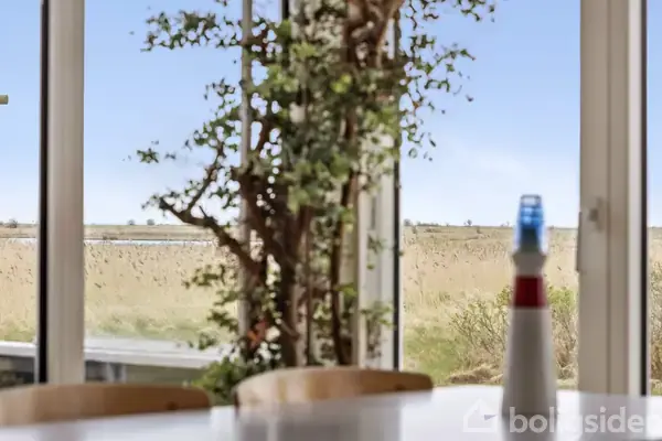 Et spisebord med en lille fyrtårn-model står foran store vinduer, der viser en udsigt over en eng eller mark med højt græs og en klar blå himmel.