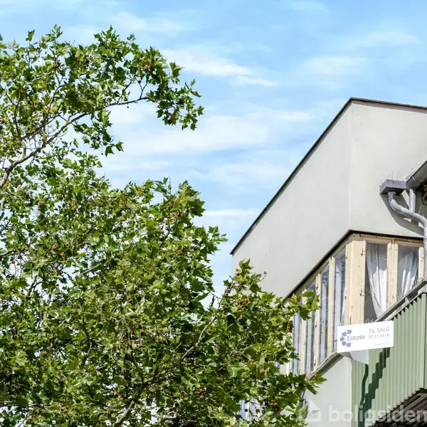 Bygning står med grene fra et træ ved siden af. En altan med hvide gardiner er synlig, og en lille hvidt skilt med teksten "Pas på dit hoved" er monteret.