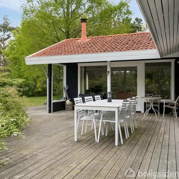 Et hvidt havebord med stole står på en træterrasse. Omkring terrassen er der grønne buske og træer. I baggrunden ses et hus med store vinduer og et rødt tegltag.