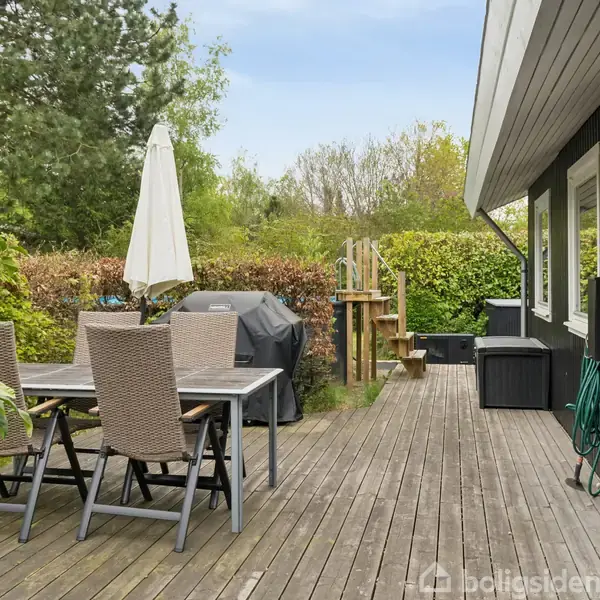 Havebord med fire stole står på en træterrasse ved siden af en grill. En hvid parasol er slået op. Området er omgivet af grønne buske og træer.