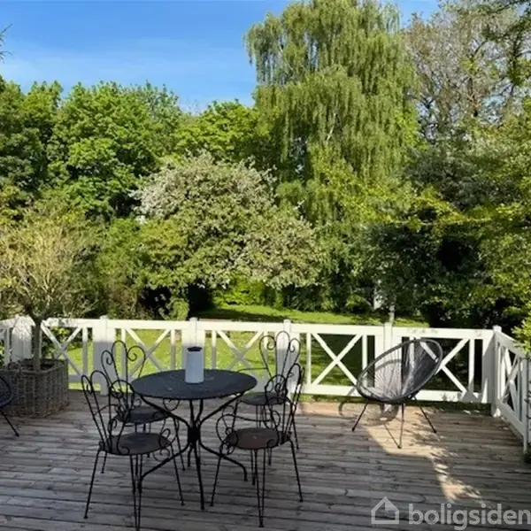 Bord med fire stole står på en terrasse, omgivet af et hvidt stakit. Terrassen har udsigt til en frodig have med forskellige træer og buske under en blå himmel.