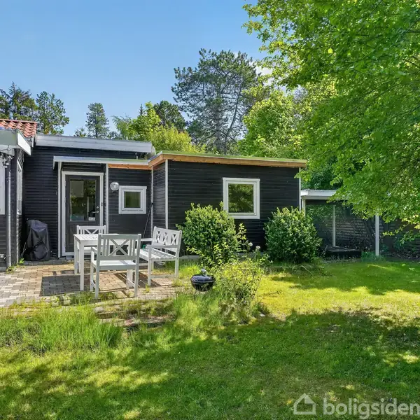Sortmalet hus står stille omgivet af frodig have og græsplæne. Et udendørs bord med stole står på en stenbelagt terrasse foran huset. Træer og buske omgiver scenen.