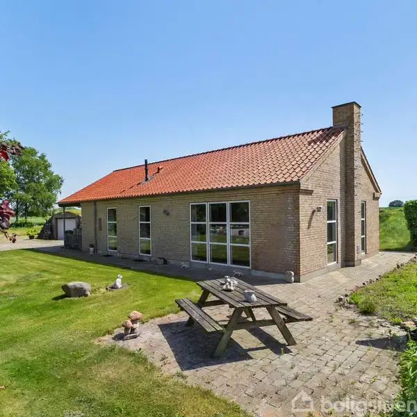 Hus med rødt tag og store vinduer står stille i en have med græsplæne og træer. Foran huset er et picnicbord på en stenterrasse.