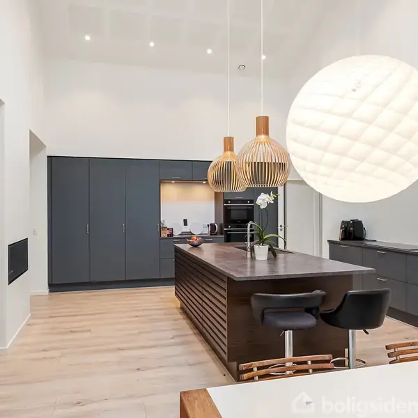 Køkken med moderne design, køkkenø med barstole og sorte skabe. Store, runde lamper hænger over øen, mens lyse trægulve strækker sig gennem rummet.