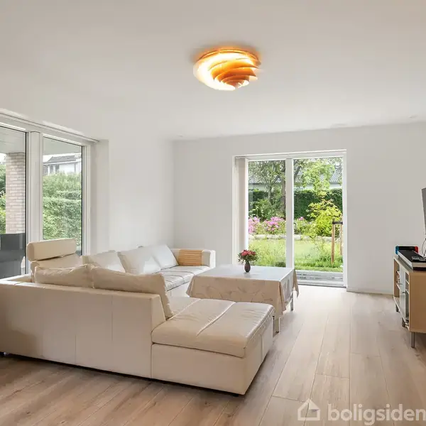 En hvid sofa står i en moderne stue med lyst trægulv. Et sofabord er foran sofaen. Et stort vindue og en dør åbner ud til en have med grønne buske.