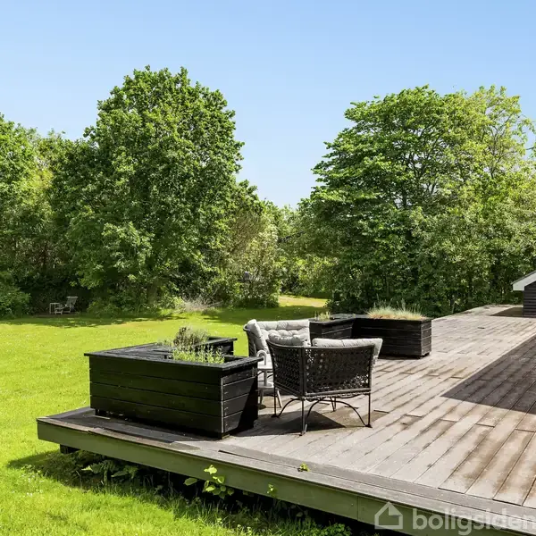 Træterrasse med siddemøbler og plantekasser. Terrassen er del af et hus, omgivet af en stor græsplæne og tætte grønne træer under en klar blå himmel.
