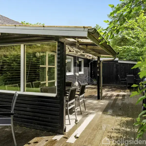 Sort træhus med store vinduer står stille. Omkring huset er en stor træterrasse med havemøbler og grønne buske. Vejret er klart med blå himmel.