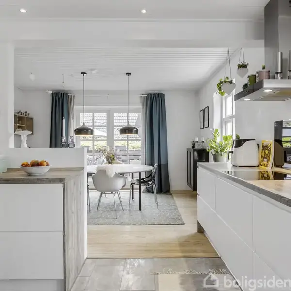 Et moderne køkken er udstyret med hvide skabe og en træbordplade, ved siden af en åben spisestue med et bord og stole. Rummet er oplyst af hængende lamper, og store vinduer lukker lys ind.