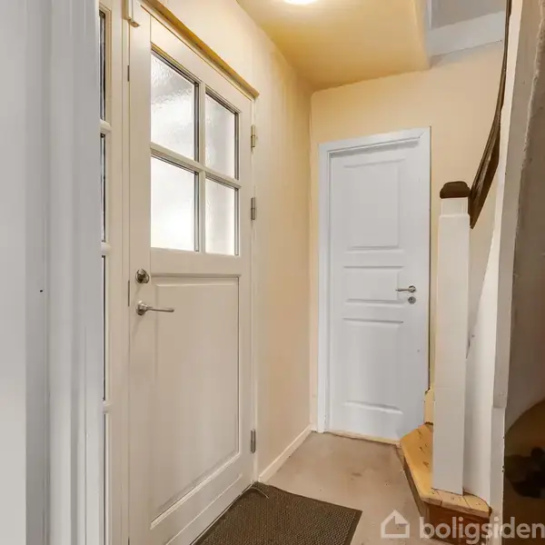 Dør fører ind i en entré med beige vægge. Til højre en trappe med mørkt gelænder. En anden dør fører videre ind i huset. Tæppe ligger på gulvet.