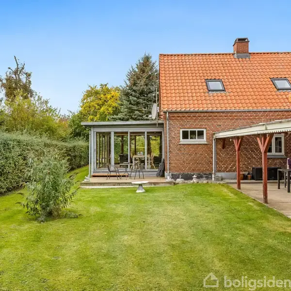 Rødt murstenshus med orangeri og overdækket terrasse, omgivet af velplejet have med grøn græsplæne og hække. Skoven ses i baggrunden under en blå himmel.