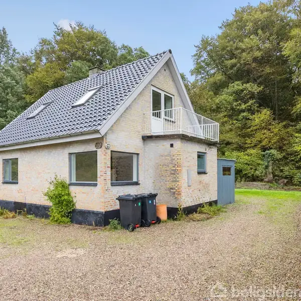 Et hvidt murstenshus med sort tag og en balkon. To skraldespande står ved siden af huset. Omgivet af en grusvej og tæt bevoksede træer i en skovlignende område.