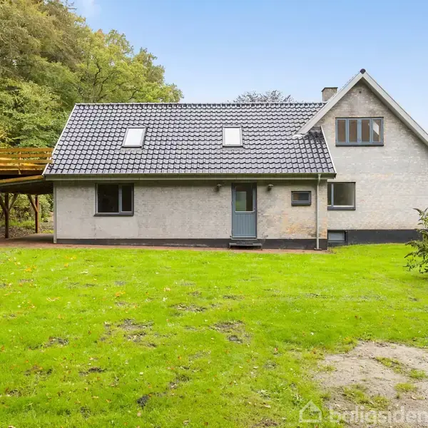 Hus med gråt tag i mursten står stille på en grøn græsplæne, omgivet af træer. En carport med fladt tag ses til venstre.