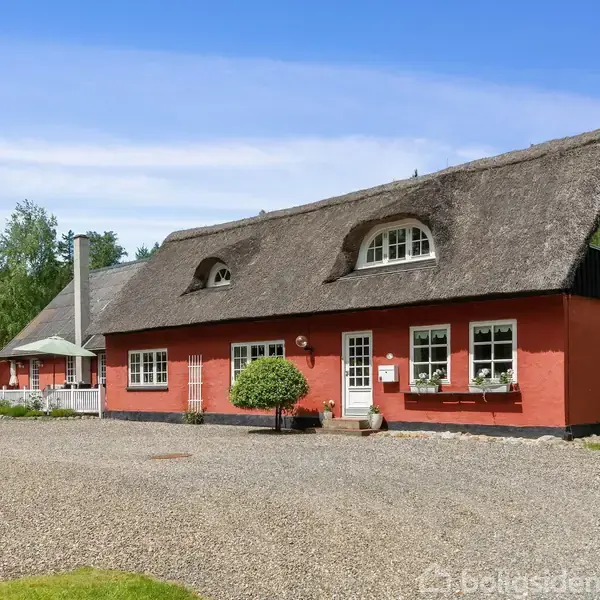 Rødt stråtækt hus med buede vinduer, omgivet af grusbelagt gårdsplads og frodig grøn beplantning i baggrunden.