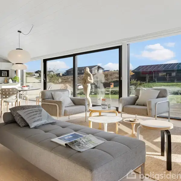 Lys stue med gråt møblement vender mod store vinduer med udsigt til have og nabolag; spisestue og køkken ses i baggrunden. Ingen tekst fundet i billedet.