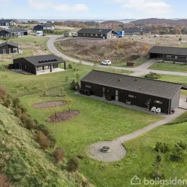Et moderne hus står omgivet af en velplejet have med stier og en lille bålplads. Omkringliggende landskab er kuperet med flere lignende huse spredt i baggrunden og en udsigt mod vandet.