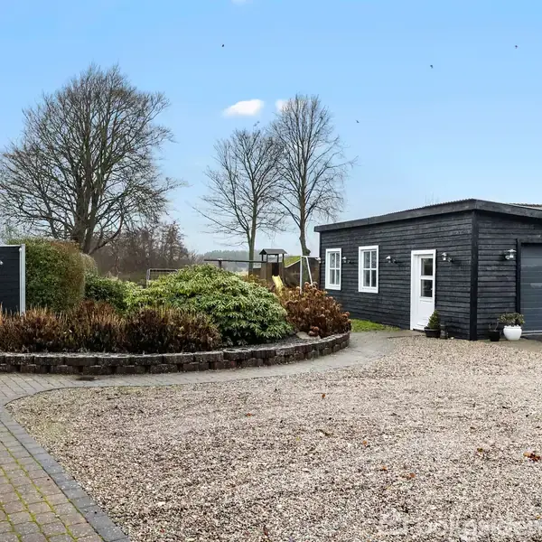 En grå garagebygning står i en gårdsplads omgivet af grus og flisebelægning, med buske og træer i baggrunden under en blå himmel.