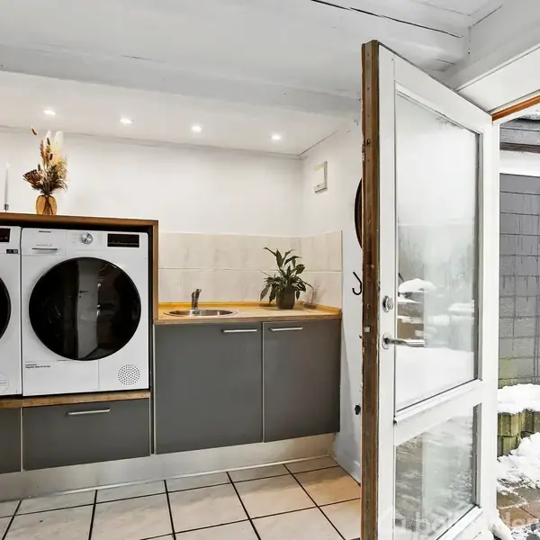 Vaskemaskine og tørretumbler stablet ved siden af en håndvask på et bord. Rummet har flisegulv, og en glasdør åbner til et snedækket område udenfor.