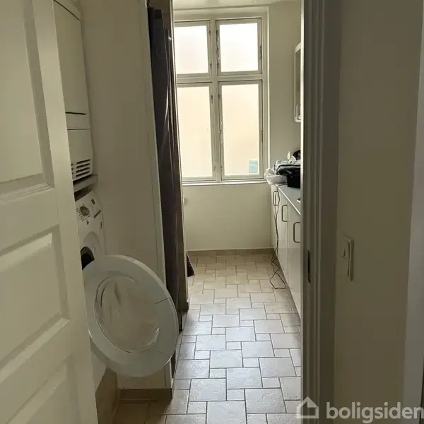En vaskemaskine med åben låge står ved siden af en tørretumbler i et smalt bryggers med klinkegulv. Til højre er skabe, og for enden er et vindue.