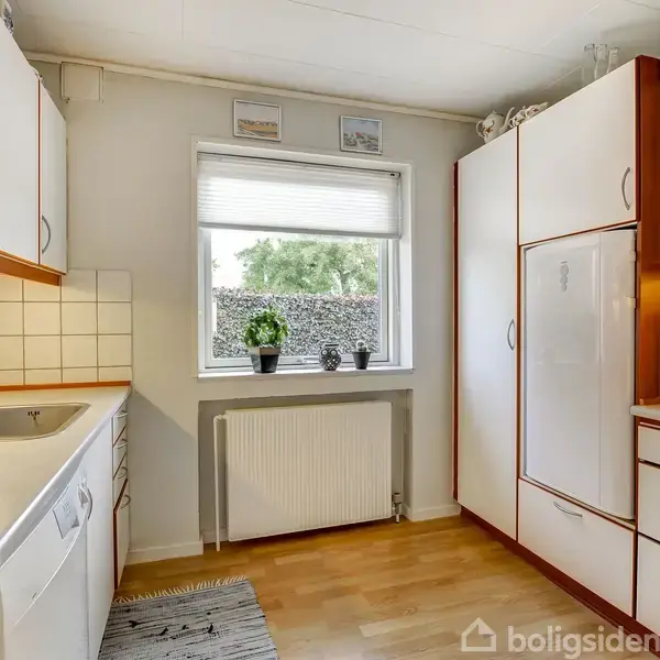 Et køkken med hvide skabe, en vask og en opvaskemaskine. Der er et vindue med planter udenfor, og en kaffemaskine står på køkkenbordet. Radiator under vinduet.