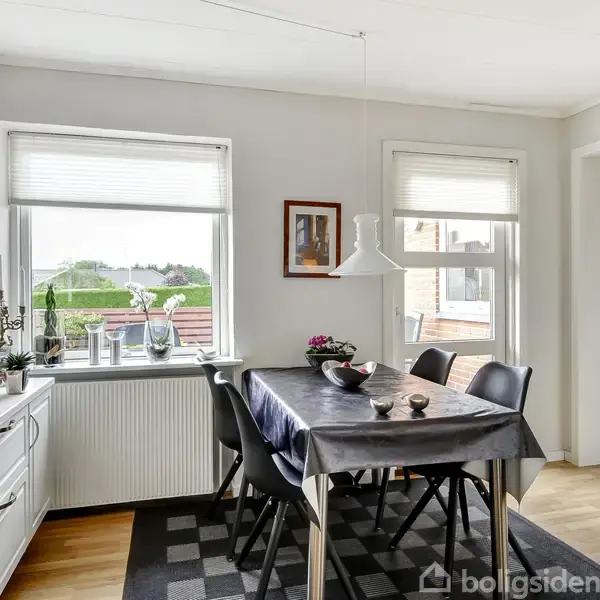 Spisebord med sorte stole står i et lyst rum ved siden af to vinduer med udsigt. Bordet er dækket med en mørk dug, og rummet har lyse trægulve og væghængt kunst.