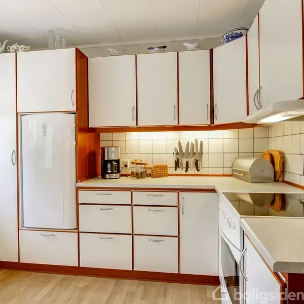 Køkkenskabe står åbne med hvide låger og røde kanter. På køkkenbordet er der en kaffemaskine, knivsæt og brødkurv. Rummet har lyse vægge og trægulv.