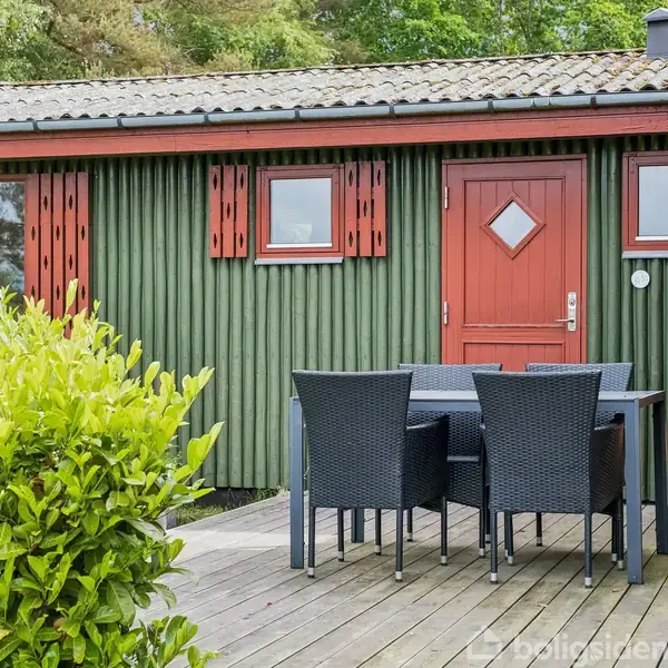 Havemøbler står på en træterrasse foran et hus med grønmalet træbeklædning og røde døre og skodder. En grøn busk står i forgrunden.