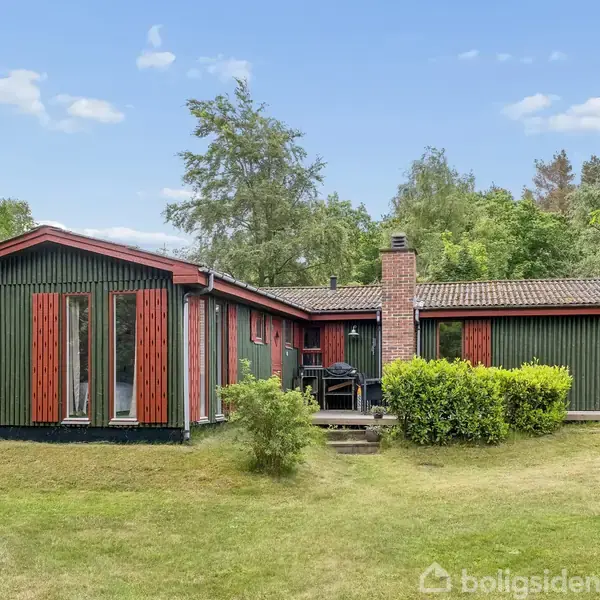 Hus i træ med grønne og røde detaljer, står stille omgivet af en græsplæne, buske og træer. Ved siden af huset ses en pejs og to stole på en terrasse.