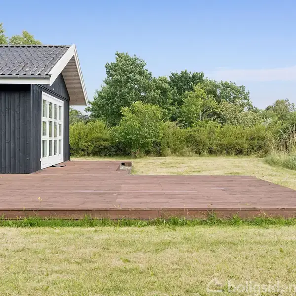 Et sort sommerhus med hvide vinduer står ved en stor træterrasse. Omkring huset er en græsplæne og grønne buske i baggrunden.