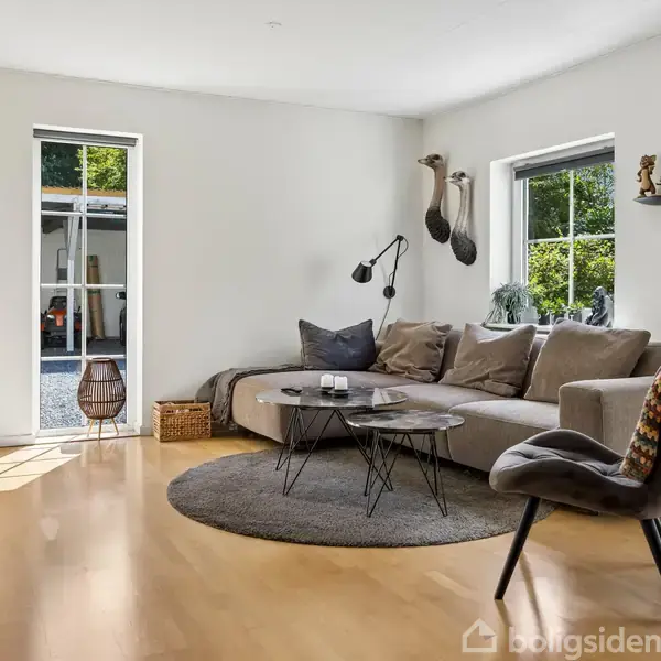 Stue med hjørnesofa fyldt med puder, sofabord, og lænestol med tæppe. Lyse vægge, store vinduer, parketgulv og loftlampe skaber en indbydende og lys atmosfære. Grønne planter pynter.