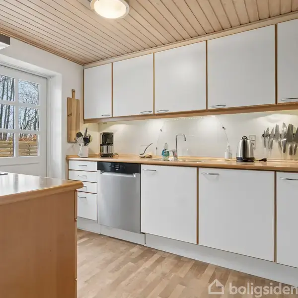 Moderne køkken med hvide skabe og indbygget opvaskemaskine. Bordpladen har køkkenredskaber og en elkedel. Et vindue til venstre lader dagslys ind, og træloftet skaber hygge.