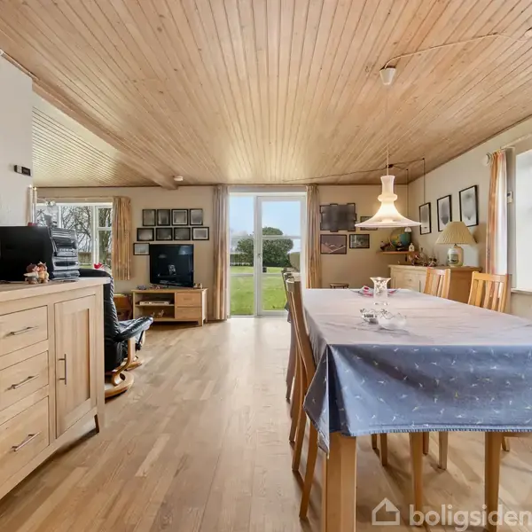 Spisebord dækket med blå dug står i midten af rummet. Træmøbler og hængelampe omgivet af trævægge og loft. Vinduespartier viser en udendørs have. Ingen tekst er synlig.