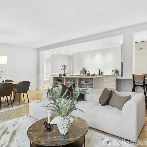 En stue i moderne stil viser en hvid sofa og sofabord med dekorative planter. Køkkenet i baggrunden har lyse skabe og en ø med barstole. Spisebordet står til venstre.