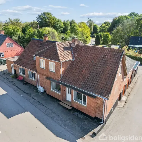 Et rødt murstenshus med skråt tag står ved vejen omgivet af andre huse og grønne træer under en blå himmel.
