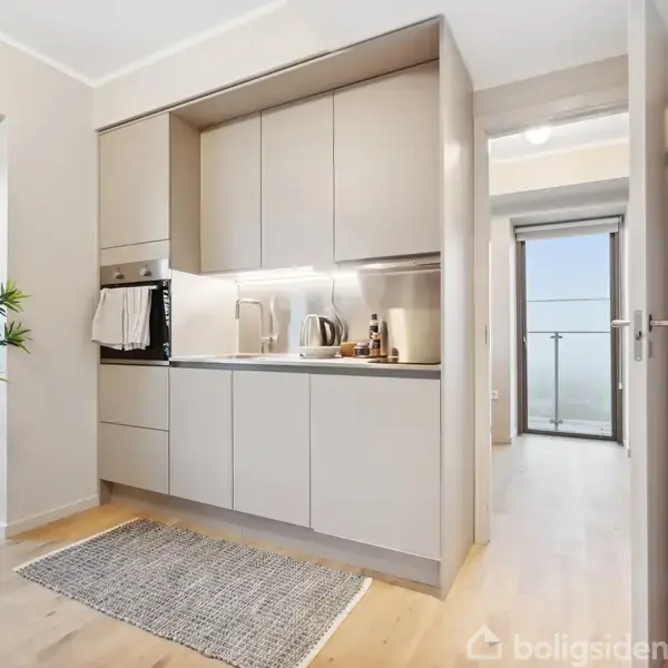 Et kompakt køkken med moderne, lyse skabe og indbygget ovn. En plante står ved siden af et vindue. En dør fører til en lys gang med glasdør i baggrunden.