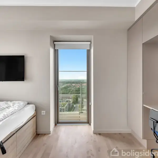 Soveværelse med seng til venstre, fladskærms-TV på væggen, skrivebord og stol til højre. Glasdør i midten fører til balkon med udsigt over landskabet.