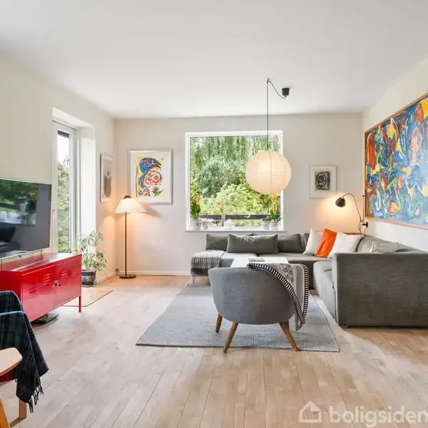 Stue indeholder en grå sofa og en grå stol, arrangeret omkring et lille bord, med farverige kunstværker på væggene. Der er en rød skænk under et TV og store vinduer, der giver naturligt lys.