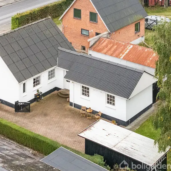 Hvid murstenshus med sort tag står stille omgivet af en gårdhave med fliser. Et par træmøbler er placeret udenfor. En vej og et murstenshus er synlige i baggrunden.