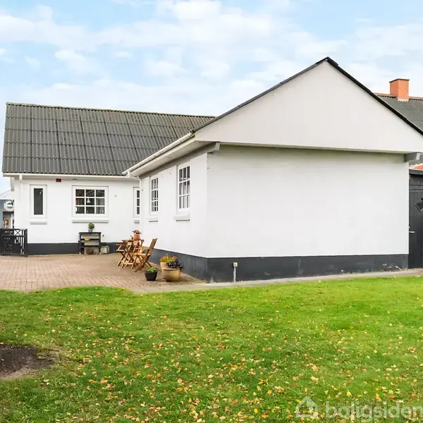 Hvidt hus med gråt tag omgivet af grøn græsplæne og flisebelagt terrasse. Havemøbler står udenfor huset ved siden af blomsterkrukker. Der er en sort bygning til venstre.