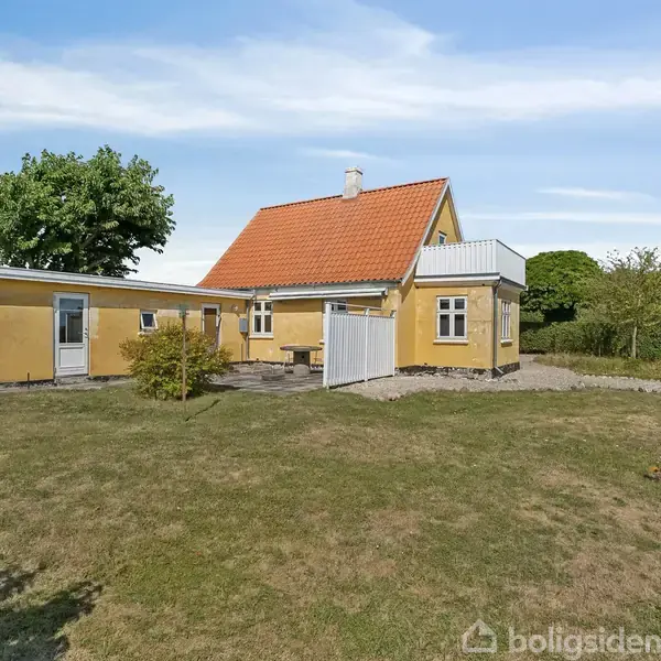 Gult hus med rødt tag står stille i en velplejet have omgivet af grønne buske og træer. En lille terrasse ses ved siden af huset under en klar blå himmel.