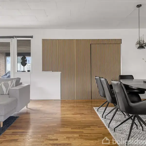 En moderne stue med grå sofa til venstre, lys trævæg i midten og spisebord med sorte stole til højre. Lyse trægulve og hvid væg udgør rummets detaljer.