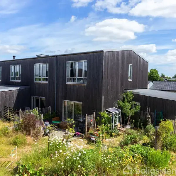 Et moderne, sort træhus med store vinduer står i en frodig have omgivet af blomsterbede og grønne områder. Huset ligger i et beboelsesområde under en blå himmel med spredte skyer.