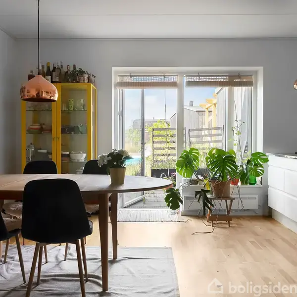 Spisebord med sorte stole står i et lyst køkken. En gul skænk med glasdøre indeholder glas og flasker. Store planter er ved vinduet, og en terrasse ses udenfor.